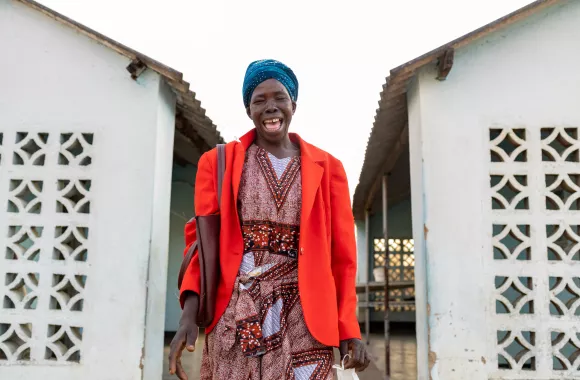 A Zimbabwean woman stands between two white buildings. She is laughing and wearing a red jacket.