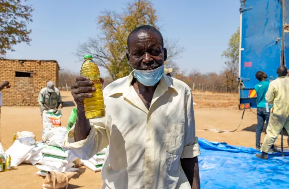 An older man with a face mask on this chin smiles for the camera and lifts up a plastic bottle of oil.