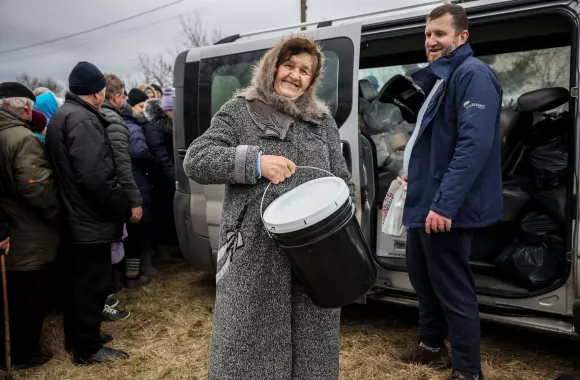 A woman in a winter coat holds up a five gallon bucket and smiles at the camera.