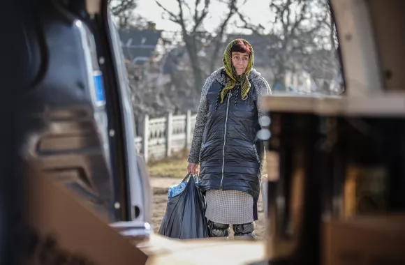 A Ukrainian woman in a winter coats holds a large bag. The view is from the perspective of someone sitting in a van