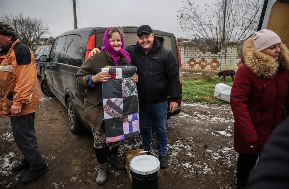 A woman and a man stand behind a van. They are wearing winter coats. The man has his arm around the woman. She is holding a folded up comforter.