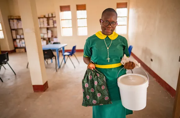 A student from South Sudan in a green and yellow uniform holds a cloth bag and a white bucket.