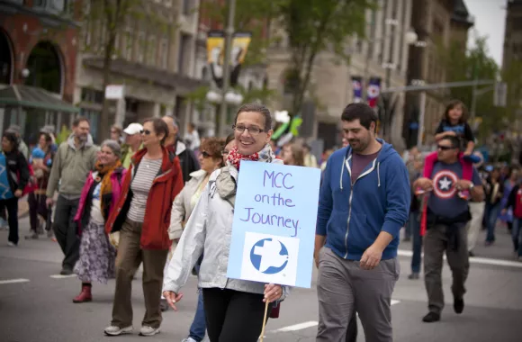 A woman walks with a large group of people during a protest. She is holding a sign that says, "MCC on the journey"