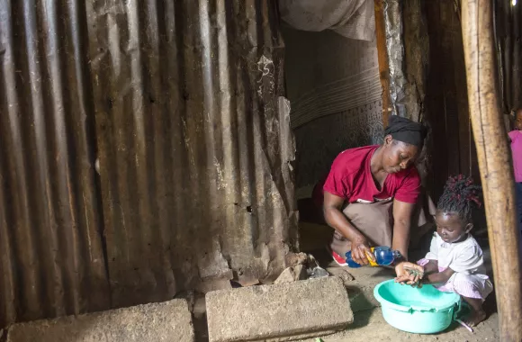 A mother helps her daughter wash her hands in a green bucket sitting outside their home