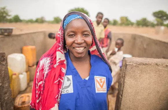 A Chadian woman in a blue shirt and red scarf covering her head smiles near a well.