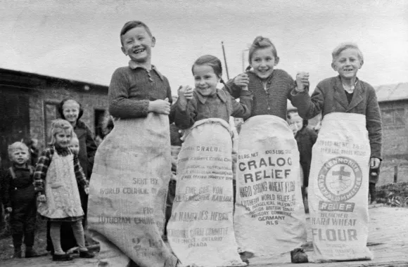 A black and white photo of four children playing in old bags used to carry MCC food supplies