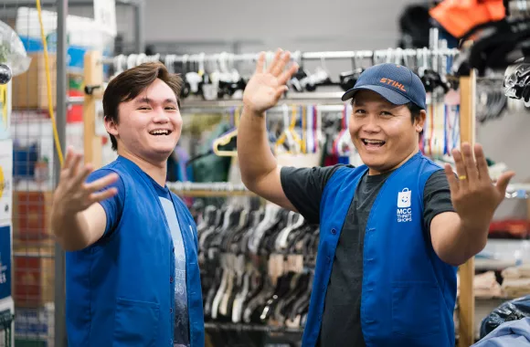 Two people wearing blue vests wave their arms in welcome. They are in a backroom of a thrift shop