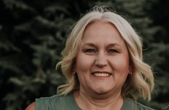 A woman with light coloured hair smiling at the camera with forestry in background.