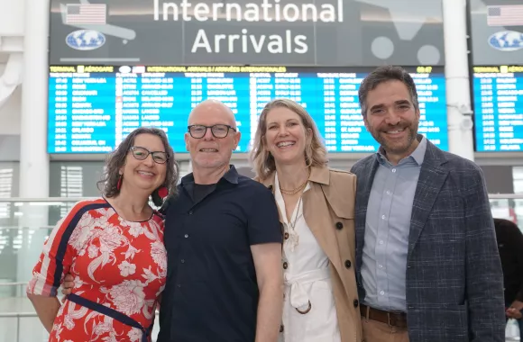 A group of four people stand in front of a sign reading international arrivals at an airport. They are smiling and facing the camera.