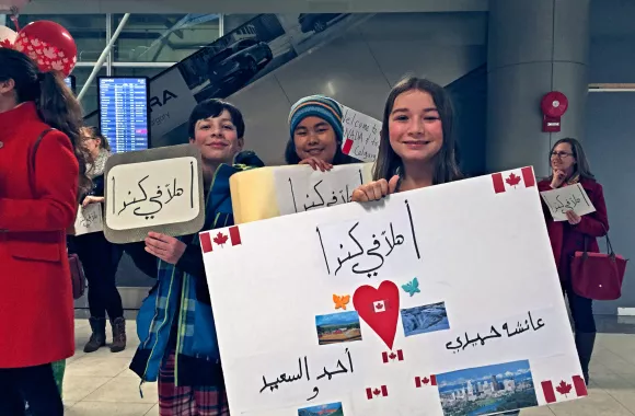 Three children welcome hold signs while waiting at an airport for a Syrian family.