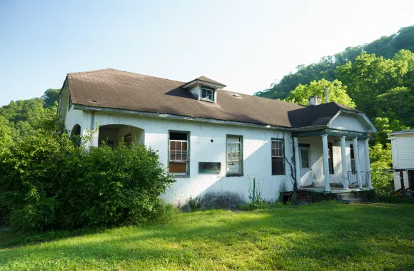 An old, neglected house with peeling white paint, surrounded by greenery, under a clear sky. It appears abandoned and in disrepair, with overgrown bushes.