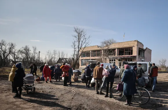 A large group of residents in a village are receiving food packages in front of a decaying building.