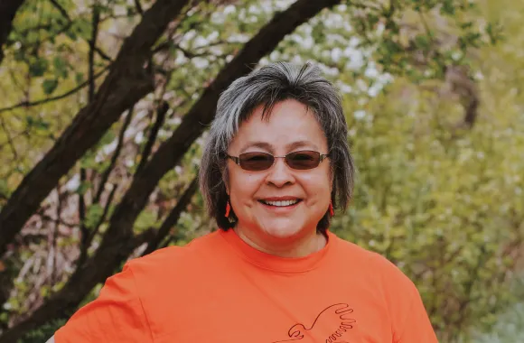 A woman wearing an orange shirt and shaded glasses stands in an outdoor area.