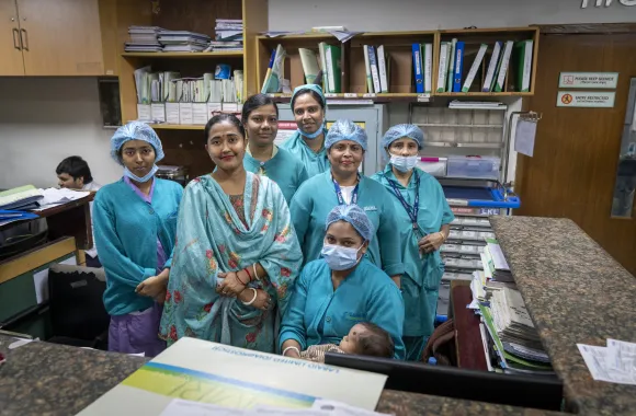 + A group of seven women in medical attire stands in an office setting, with files and equipment visible in the background.