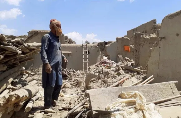 A person stands amidst the rubble and debris that was his house, with a ladder leaning against a damaged wall and remnants of structures around.