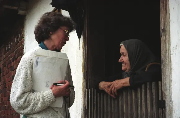 A person speaks to another, who is leaning over a gate.