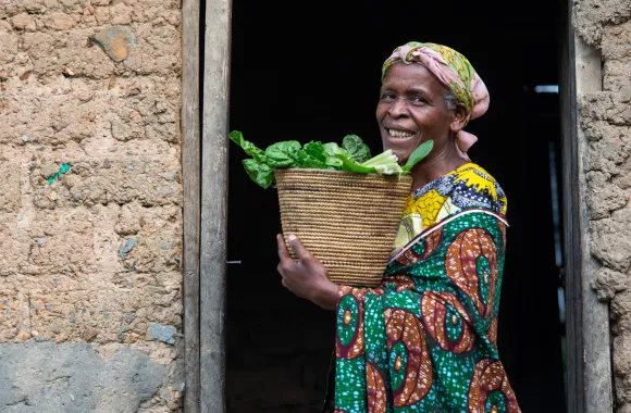 A smiling woman in colorful attire stands in a doorway holding a basket of leafy greens. The building appears to be made of mud bricks.