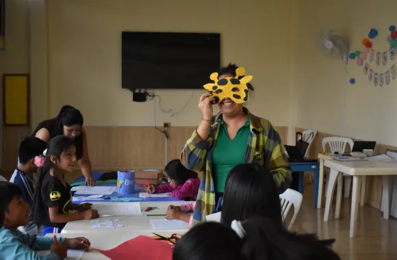 Person holding a giraffe mask in front of their face in a classroom with children engaged in arts and crafts activities.