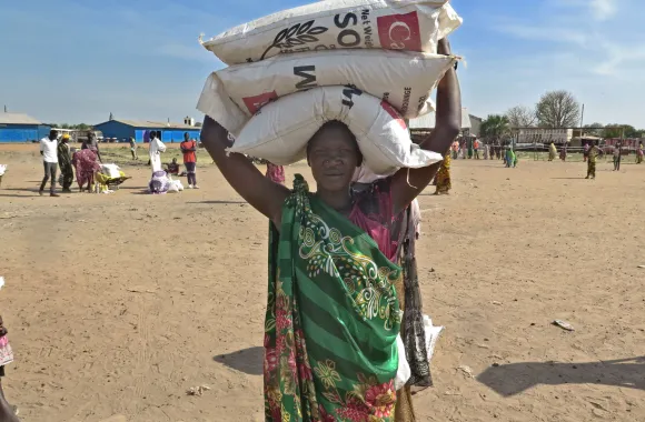 A person stands outside balancing three bags of grain on their head.