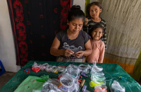 Maria Cali Tźiná behind her beadwork table with Juana Alicia Vasquez Cali (a young girl she cares for) and daughter Genesis Maria Tuit Cali (4). Maria is able to sell her beadwork with the support o