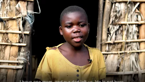 A young child stands outside her hut in the DR Congo. She is wearing a yellow shirt.