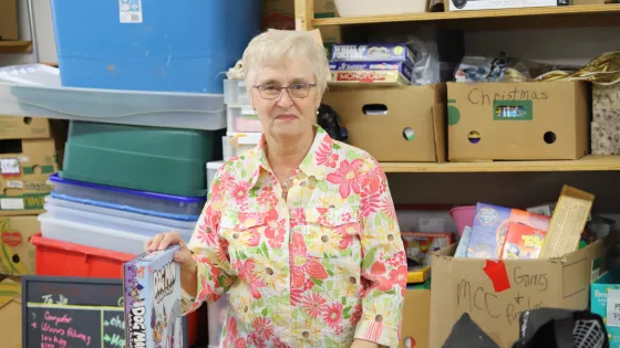 An older woman in a floral shirt stands in front of a wall of thrift shop donations