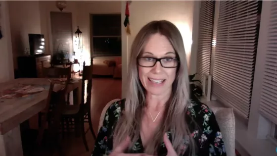 A woman with long hair and glasses speaks into the camera during a video call