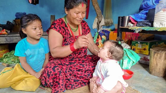 A Nepali woman sits on the ground and feeds a baby with a spoon. Another child is sitting to the left of her.