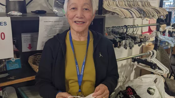 An old woman in a black jacket and green shirt smiles for the camera. She is standing in front of a rack of clothes hangers. 