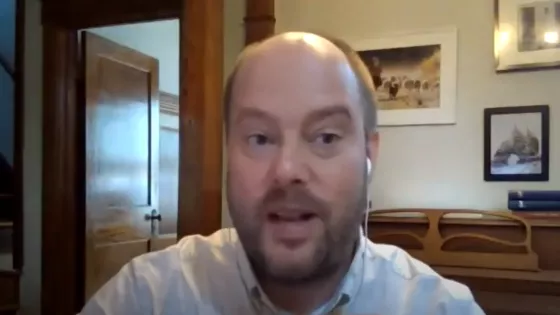 A balding man with beard and a white collared shirt speaks into camera during video call