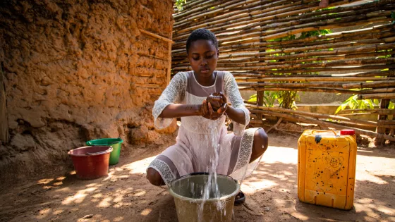 A girl in a white dress washes her hands in a bucket. She has a stream of water falling off her hands.