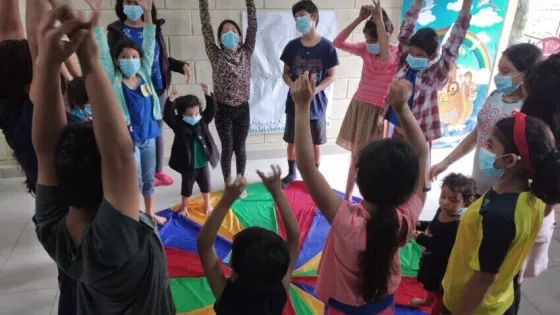 A group of children stand around a colorful parachute on the ground. They are raising their hands in the air.