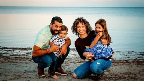 A family of four kneal together in the sand at the beach. The ocean is behind them.