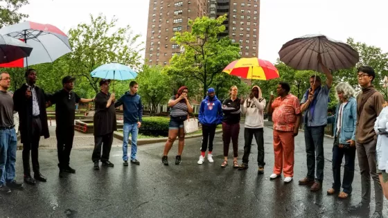A group of young adults stand on pavement in a circle. Many of them are holding umbrellas.