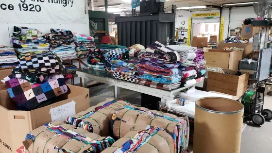 A number of piles of folded comforters sit on different surfaces in a warehouse.