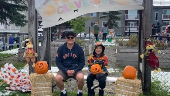 A father and son sit together on a bunch. The son is holding a carved pumpkin