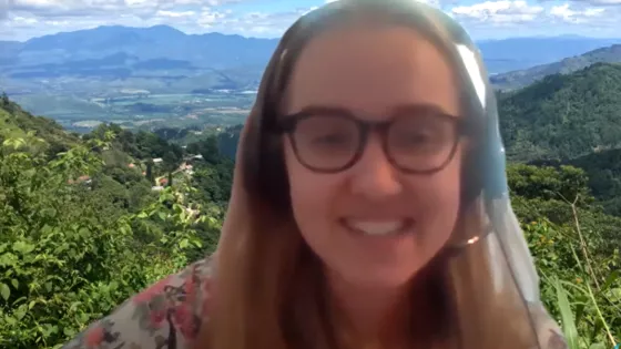 A young woman speaking during a webinar zoom call. There is a zoom background of mountains behind her.