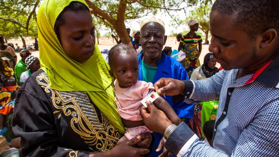 A woman holding her child while a man measures the circumference of the child's arm