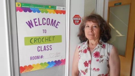 An older woman stands near a sign posted on a window that says, "Welcome to crochet class"