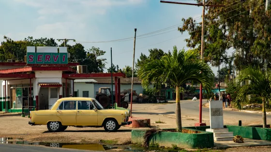 An old yellow car is parked outside of a dated gas station.