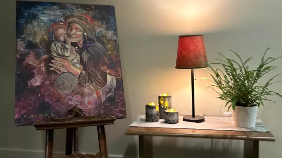 A display with art, a lamp, candles and flowers. The art is a picture of a mother holding a baby.