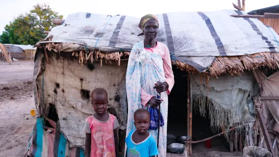 Mary Nyashin Tsief (70) stands in front of her home in Rubkona camp for people displaced by flooding and violent conflict. She is accompanied by her two grandchildren, Nyamen Iuok Ram (age 5) and Nyawich Iuok Ram (age 3), who live in a tent adjacent to hers.