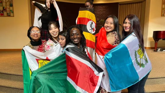 A group of smiling young adults stand posed while wrapped in flags from various nations.