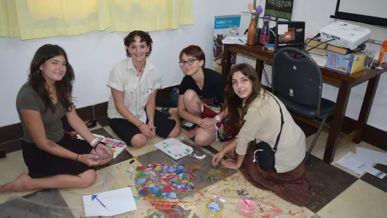 Four individuals are seated on the floor, surrounded by art supplies and colorful paper cutouts. They appear engaged in a creative activity, smiling at the camera in a room with a casual setting.