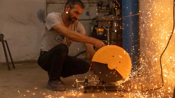 A man is crouching while using an angle grinder on metal, causing bright sparks to fly in a workshop setting.