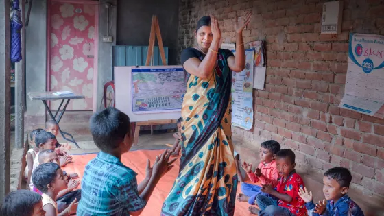 A person, surrounded by clapping children stands with her arms raised in a clapping motion.