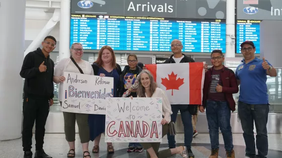 A group of people stand in an airport with a Canadian flag and signs that read "Welcome to Canada"