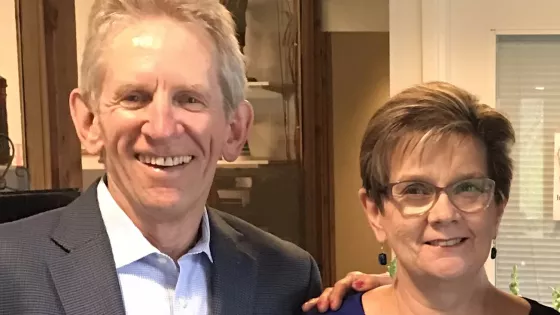 The photograph features two smiling individuals standing close together in a well-lit indoor setting. One person is wearing a suit, while the other is dressed in a stylish top. They appear to be enjoying a friendly moment, with a warm and inviting atmosphere around them.