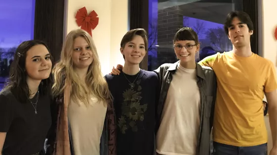 The photograph features five young individuals standing together, smiling and posing in a casual setting, with a festive decoration visible in the background, suggesting a celebratory atmosphere. Their expressions convey warmth and camaraderie.