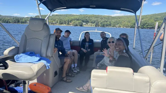 Seven people sit together on a boat in a like with green hills in the distance.